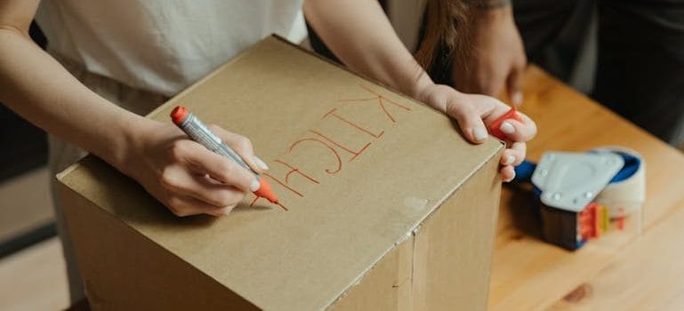 Woman in White Shirt Holding Red Pen Writing on Brown Cardboard Box